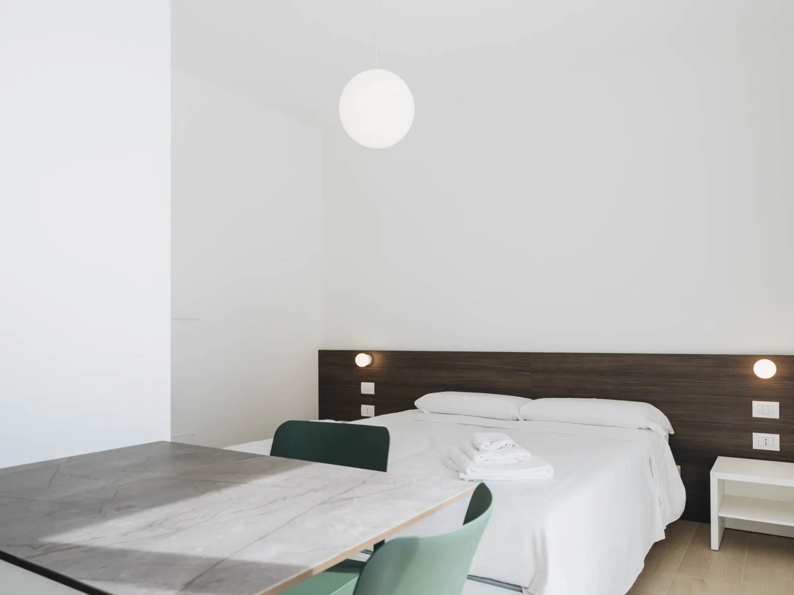 Letto matrimoniale con testiera in legno scuro, comodino bianco e tavolo pranzo in primo piano – appartamento T-07 Ca' Sette Treviso
